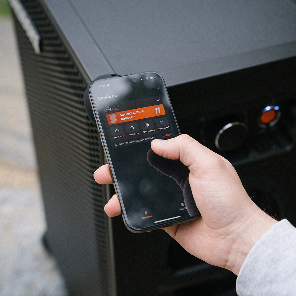 A person holds a smartphone in their hand, displaying the SOUNDBOKS app with options for controlling connected devices. Behind the phone is a large SOUNDBOKS 4 party speaker, with visible control buttons.